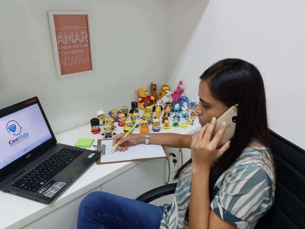 mulher sentado em frente a notebook anotando algo em caderno e falando ao telefone